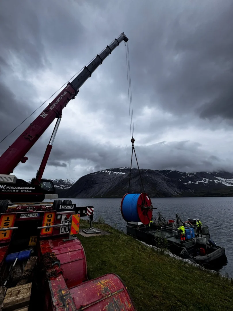 Mobile crane lifting a cable drum onto a work barge at the lake shore